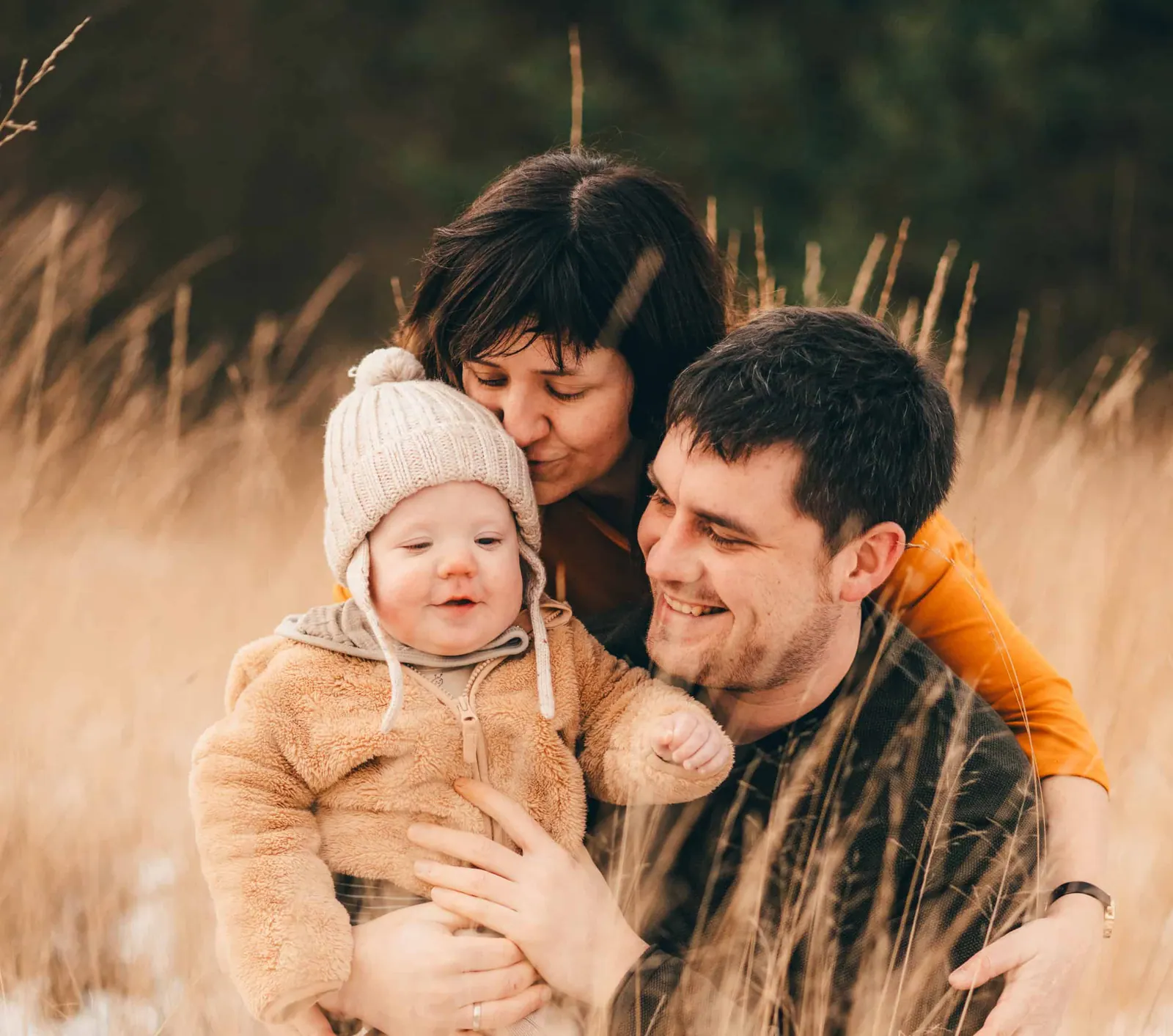 Jong gezin met een baby in een wollen mutsje lacht samen in het hoge gras