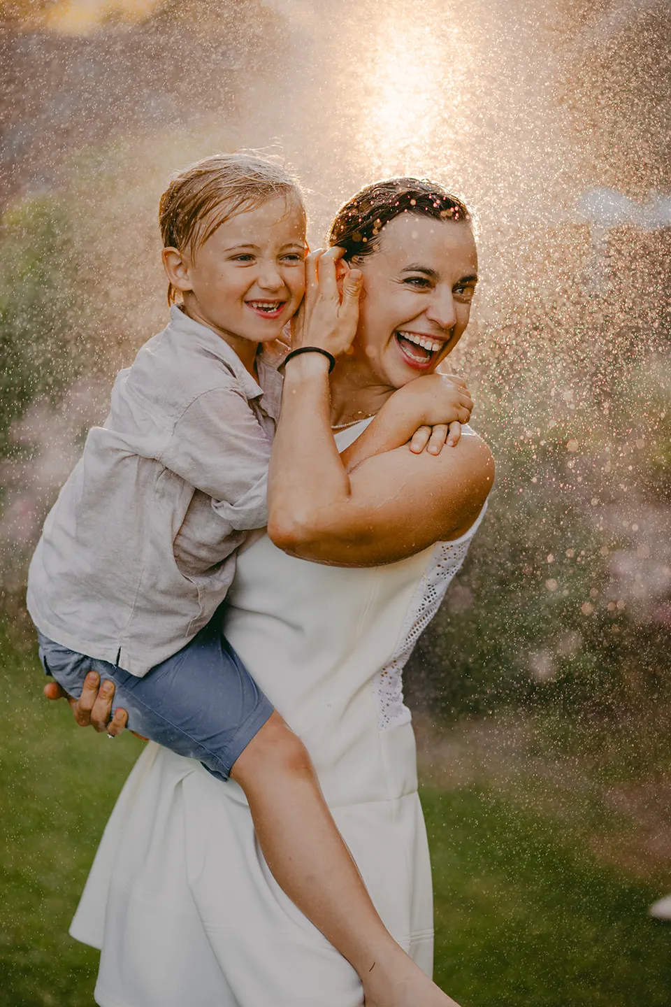 Mama tilt haar zoon op in de glinsterende waterspray van de tuinslang